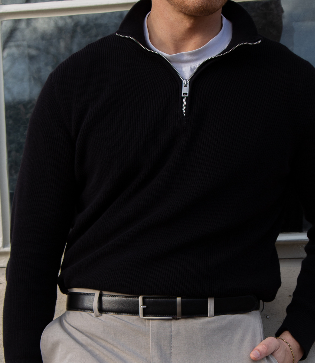 Een model leunt casual tegen een muur en draagt de zwarte Buckle Up-riem, wat zorgt voor een moderne en stoere uitstraling.